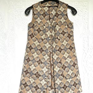 May include: A sleeveless vest featuring a repeating diamond pattern in shades of tan, brown, cream, and muted blue. The garment has a V-neckline, a front button closure with metallic snaps, and two patch pockets on the lower front section.