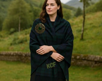 May include: A dark green and navy blue plaid shawl with a decorative gold brooch featuring a green stone. The shawl has a fringed edge and the initials "KCF" are embroidered on the front. The background is a blurred outdoor scene.