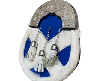 Puede incluir: Un pin de kilt azul y blanco con un diseño de metal plateado y ribete de piel blanca. El pin tiene tres borlas blancas que cuelgan.
