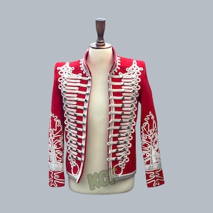 May include: A red military-style jacket with white and silver decorative trim. The jacket features a high collar, multiple rows of silver buttons, and ornate embroidery on the sleeves and shoulders. The jacket is displayed on a mannequin.