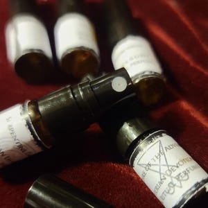 May include: A collection of small amber glass perfume spray vials with black caps and white labels. One label features a pentagram symbol and the text "NATURE OF THE ADVENT" and "RITUAL PERFUME," while another shows "SERPENTINE."
