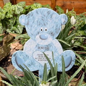 Op de afbeelding: Een blauwe teddybeer-tuinpaal met een hartvormige boodschap die luidt: "Je bent misschien niet in onze armen, maar je zult voor altijd in ons hart zijn." De paal staat in een bed van groene planten met witte bloemen.