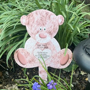 Op de afbeelding: Een roze teddybeer-vormige tuinpaal met de tekst "BABY THAPA 07-03-2024" en een hartvormige inscriptie: "YOU MAY NOT BE IN OUR ARMS BUT YOU BE FOREVER IN OUR HEARTS". De paal staat in een tuin.