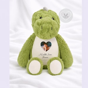 May include: A plush green dinosaur toy with a soft, textured exterior. The center of the chest features a white patch with a heart-shaped photograph of a person and the printed text A Cuddle from Heaven followed by a small heart icon.