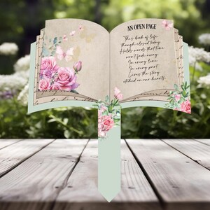 May include: A decorative garden stake shaped like an open book. The book features pink roses, butterflies, and the text "AN OPEN PAGE." The stake has a light green base and is set on a wooden surface.