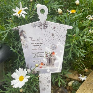 May include: A white garden stake with a decorative top, featuring a bow and snowflake design. The stake has the text "Those we Love...every day" with an illustration of robins and winter boots. The stake is surrounded by green foliage and white daisies.
