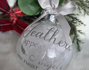 May include: Clear glass Christmas ornament filled with white feathers and the text "Feathers appear when Angels are near" in silver script. The ornament is tied with a silver satin bow.