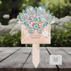 Può includere: Un paletto da giardino commemorativo con una composizione floreale a forma di busta. La composizione comprende fiori blu, rosa e pesca. Il testo recita "IN LOVING MEMORY" e "Mum". Il paletto è rosa chiaro con accenti glitterati dorati.