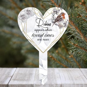 May include: A heart-shaped garden stake featuring a white background with gray floral illustrations, a gold geometric frame, and a robin bird. The text reads, Robins appear when loved ones are near, printed in black script and serif lettering.