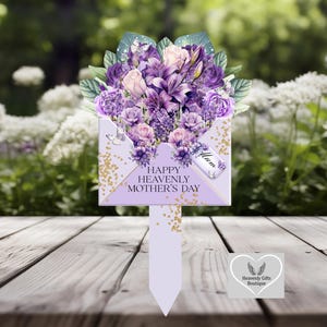 Op de afbeelding: Een lavendelkleurige Moederdag-tuinsteker met een boeket paarse en roze bloemen die uit een decoratieve envelop komen. De steker heeft de tekst "Happy Heavenly Mother's Day" en een label met "Mum".