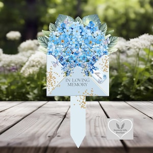 Può includere: Un paletto da giardino commemorativo azzurro con un bouquet di fiori blu e farfalle. Il paletto ha la forma di una busta con le parole "IN LOVING MEMORY" e un'etichetta con la scritta "Mum". Accenti di glitter dorati.