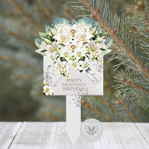 Może przedstawiać: Pamiątkowy palik ogrodowy z motywem białej koperty zwieńczony bukietem białych lilii i zielonych liści. Na kopercie widnieje napis "HAPPY HEAVENLY BIRTHDAY" oraz mała zawieszka ze słowem "Mum" i czarną kokardką.