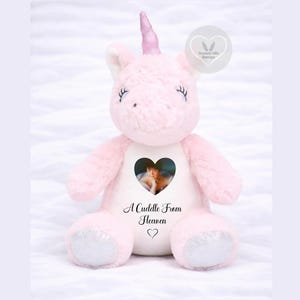 May include: A soft, light pink plush unicorn toy with a glittery pink horn and silver glittery foot pads. The chest features a heart-shaped photo of a person, the text "A Cuddle From Heaven," and a small heart icon below the text.