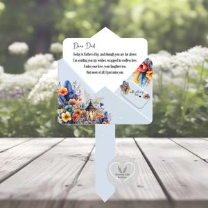 Op de afbeelding: Een lichtblauwe herdenkingspaal voor Vaderdag met een bloemenontwerp en de tekst "Lieve papa". De paal heeft een lantaarn en een klein label met de tekst "met al mijn liefde". De onderkant van de paal heeft een hartvormig logo met "Heavenly Gifts Boutique".