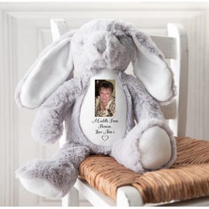 May include: A grey plush bunny toy with long ears, sitting on a chair. The bunny has a white patch on its chest with a photo and the text "A Cuddle From Heaven, Love Nan x". A soft, cuddly toy.