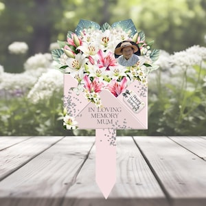 Pode incluir: Uma estaca de jardim comemorativa com design de envelope rosa e o texto IN LOVING MEMORY MUM. O topo exibe um buquê de lírios brancos e rosas, uma foto circular de uma pessoa usando chapéu e uma etiqueta escrita Happy Heavenly Birthday.