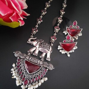 May include: A silver necklace with a red stone pendant featuring an elephant design. The necklace has a chain with small silver beads and red stones. The pendant has a detailed elephant design with a red stone in the centre. The necklace comes with a pair of matching earrings with red stones.