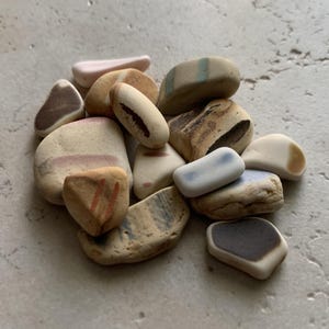 May include: A collection of smooth, colorful stones in various shapes and sizes. The stones feature a palette of beige, brown, pink, and blue hues, with some displaying striped or speckled patterns. The arrangement suggests a natural, organic theme.