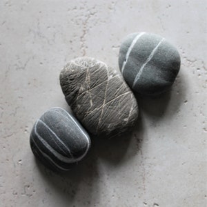 May include: Three smooth, gray stones with white lines. The stones are arranged in a triangular shape on a light gray background.