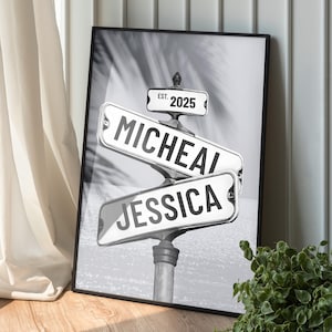 May include: Black and white framed print featuring street signs with the names "Micheal" and "Jessica". A smaller sign reads "Est. 2025". The background is a blurred image of palm trees. The print is leaning against a white wall.