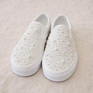 May include: A pair of white slip-on sneakers embellished with numerous faux pearls of varying sizes. The shoes have a white rubber sole and a clean, minimalist design, suitable for special occasions or everyday wear.