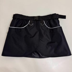 May include: Black skirt with a white trim and a black belt. The skirt has two pockets on the front and two metal rings on the waistband.