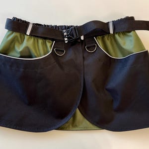 Può includere: Gonna a portafoglio nera e verde oliva con cintura nera e accessori in metallo color argento. La gonna ha due tasche anteriori con finiture riflettenti. La cintura ha una fibbia in plastica e strisce riflettenti.