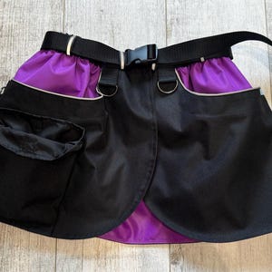 May include: Black and purple apron with a black belt and silver buckle. The apron has two pockets and a purple lining. The belt is adjustable. The apron is designed for gardening or crafting.