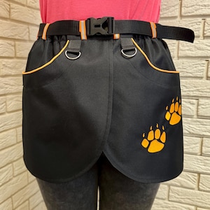 May include: Black apron with orange trim and paw print design. Features a black belt with a buckle and orange reflective stripes. Includes two D-rings and two pockets. The apron is designed to be worn around the waist.