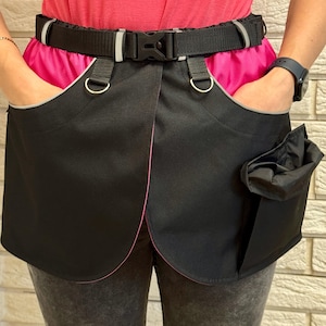 May include: Black waist apron with a pink interior and a black belt. The apron has two large pockets with grey trim and a small pouch on the right side. The belt has a black buckle and reflective strips.