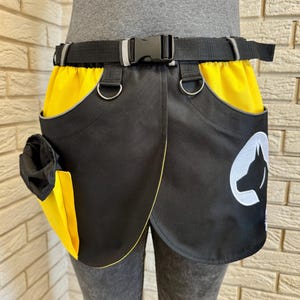 May include: Black and yellow dog training treat bag with a black belt and buckle. The bag features a dog silhouette logo and D-rings for accessories. The bag has a pocket for treats and a yellow lining.