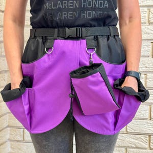 May include: A purple and black utility apron featuring a black adjustable waist belt with a plastic buckle. The apron includes two large side pockets with black trim and a detachable purple and black pouch secured by a metal clip and D-ring.
