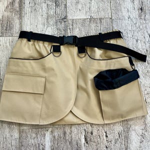 May include: A tan skirt with black detailing and a black belt. The skirt has two front pockets, black D-rings, and a wrap-around design. The black belt has a plastic buckle.