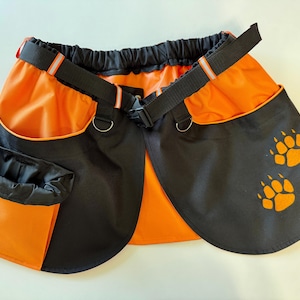 May include: An orange and black apron-style dog treat bag with a black adjustable belt and D-rings. The apron has two pockets and a pouch. Two orange paw prints are printed on the black side panels.
