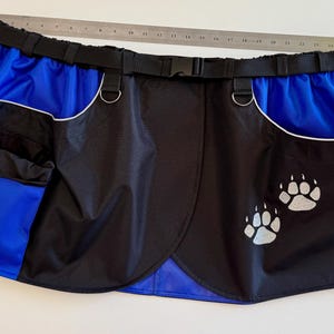May include: Black and blue dog training apron with a black belt and silver hardware. The apron has two pockets, a blue interior, and white paw prints. A metal ruler is placed across the top of the apron.
