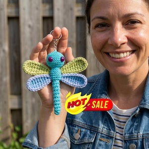May include: A crocheted dragonfly keychain featuring a bright turquoise body, two light green upper wings, and two light blue lower wings. The dragonfly has two dark purple bead eyes and is attached to a silver-toned metal keyring.