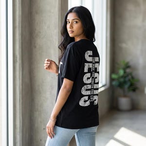 May include: Black t-shirt with the word "JESUS" printed vertically in distressed white font on the back. The text "THE WAY THE TRUTH THE LIFE" is printed vertically next to the word "JESUS". A white cross is visible on the front.