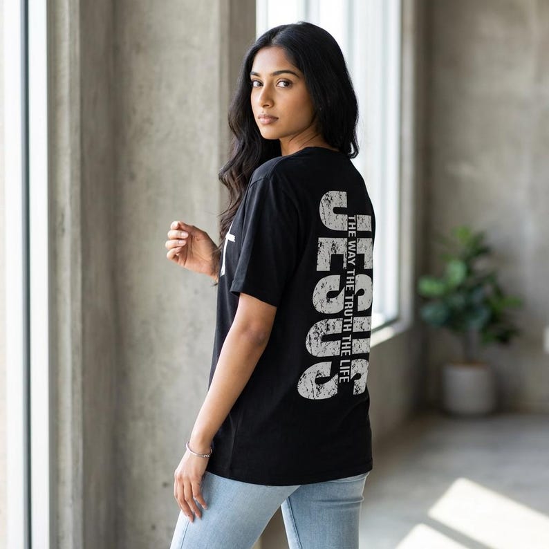 May include: Black t-shirt with the word "JESUS" printed vertically in distressed white font on the back. The text "THE WAY THE TRUTH THE LIFE" is printed vertically next to the word "JESUS". A white cross is visible on the front.