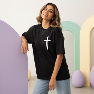 May include: Black t-shirt with a white cross graphic. The short-sleeved shirt is made of a soft material. The cross is a simple, brushstroke design. The shirt is paired with blue jeans.