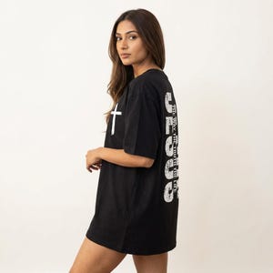 May include: Black oversized t-shirt with a white cross graphic on the front and the word "JESUS" printed vertically on the back. The text "THE WAY, THE TRUTH, THE LIFE" is printed vertically on the back. The shirt is made of a soft material.