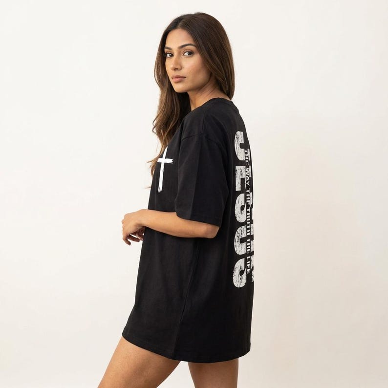 May include: Black oversized t-shirt with a white cross graphic on the front and the word "JESUS" printed vertically on the back. The text "THE WAY, THE TRUTH, THE LIFE" is printed vertically on the back. The shirt is made of a soft material.