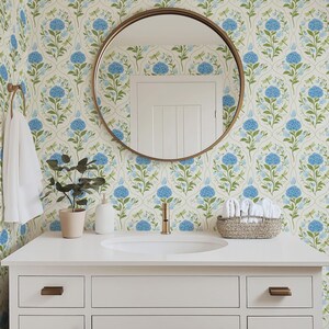 May include: A bathroom vanity with a white countertop, a round mirror, and a floral wallpaper featuring blue hydrangeas and green leaves. A small potted plant, a soap dispenser, and a woven basket with white towels are on the countertop. The faucet and hardware are gold.