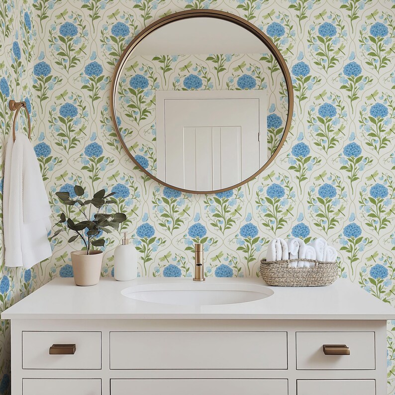 May include: A bathroom vanity with a white countertop, a round mirror, and a floral wallpaper featuring blue hydrangeas and green leaves. A small potted plant, a soap dispenser, and a woven basket with white towels are on the countertop. The faucet and hardware are gold.