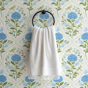 Close-up of a white hand towel hanging on an oil-rubbed brass ring, with blue hydrangea floral wallpaper in the background featuring butterflies and dragonflies on a white background.