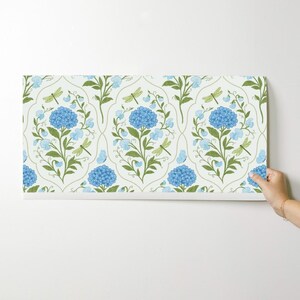 May include: A roll of wallpaper featuring a repeating pattern of blue hydrangea flowers, green leaves, and dragonflies on a cream background. The design includes light blue butterflies and a stylized floral motif.