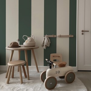 May include: A round wooden table with a matching stool, a beige woven basket, a brown hat, and a cream-colored ride-on toy car labeled with the number 10. The room features vertical dark green and beige striped wallpaper and light wood wall hooks.