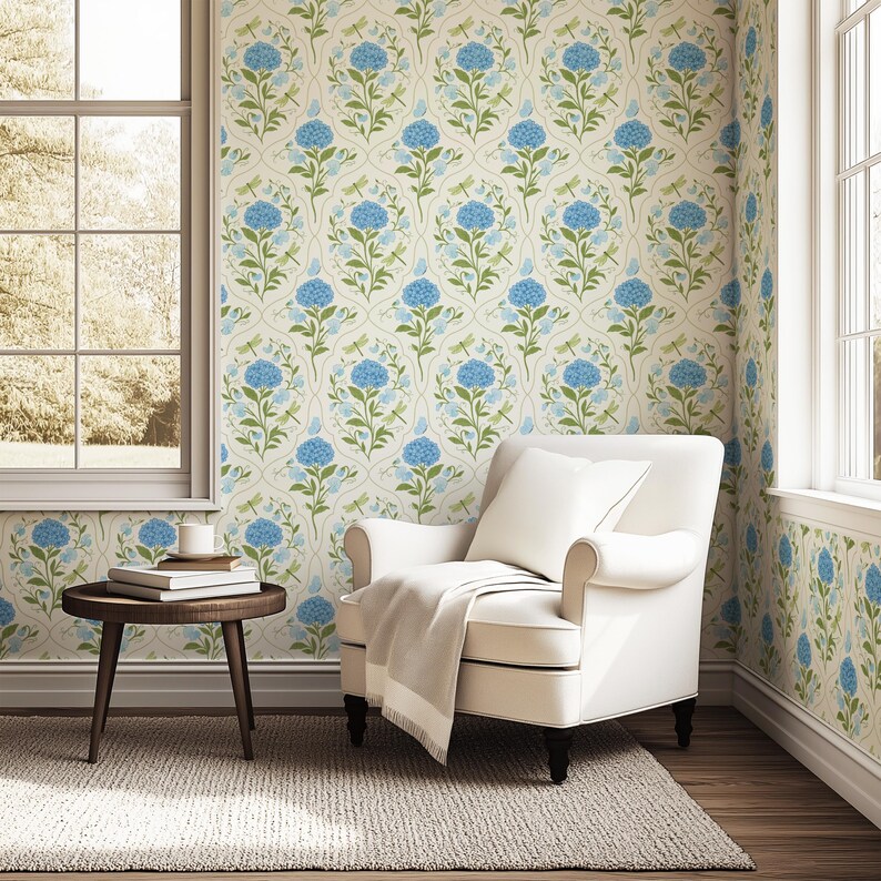 May include: A cozy room featuring floral wallpaper with blue hydrangea blooms and green leaves. A white armchair with a blanket and pillow sits on a neutral rug. A small wooden side table holds books and a mug.