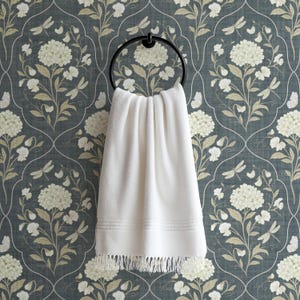 May include: A white, fringed hand towel hangs from a black metal ring. The towel has a textured surface and decorative lines near the bottom. The background is a floral wallpaper in shades of blue, cream, and beige.