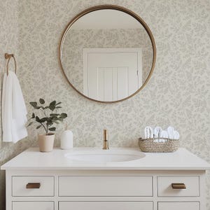 May include: A round mirror with a thin bronze frame hangs above a white bathroom vanity with a sink, a faucet, a potted plant, a white soap dispenser, and a woven basket containing white towels. The wall features a light floral wallpaper pattern.