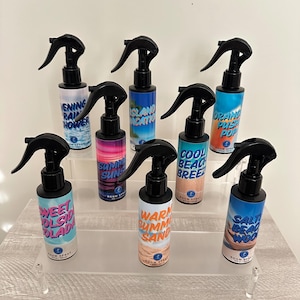 May include: A collection of room sprays with black spray nozzles and white labels. Each label features a different summer-themed scent name, such as "Evening Rain Shower," "Island Vacation," and "Orange Push Pop." The bottles are displayed on a clear, tiered shelf.